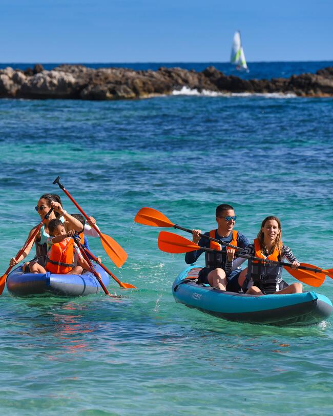 Zwei Kajaks im klaren blauen Wasser, Menschen mit orangefarbenen Paddeln und Schwimmwesten, felsige Küste im Hintergrund.