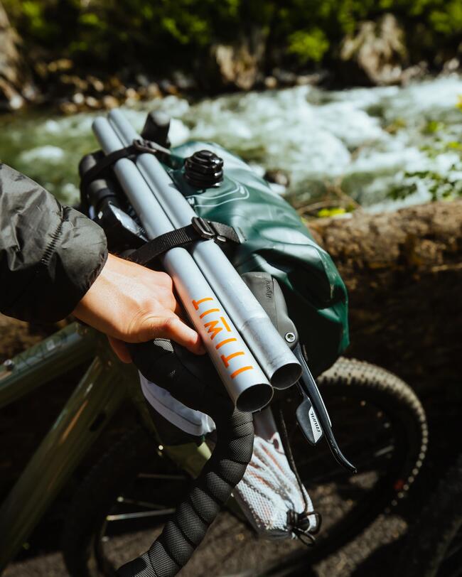 Ein zerlegtes Paddel und ein verpacktes Raftingboot sind an den Lenker eines Fahrrades geschnallt. Im Hintergrund ein Fluss