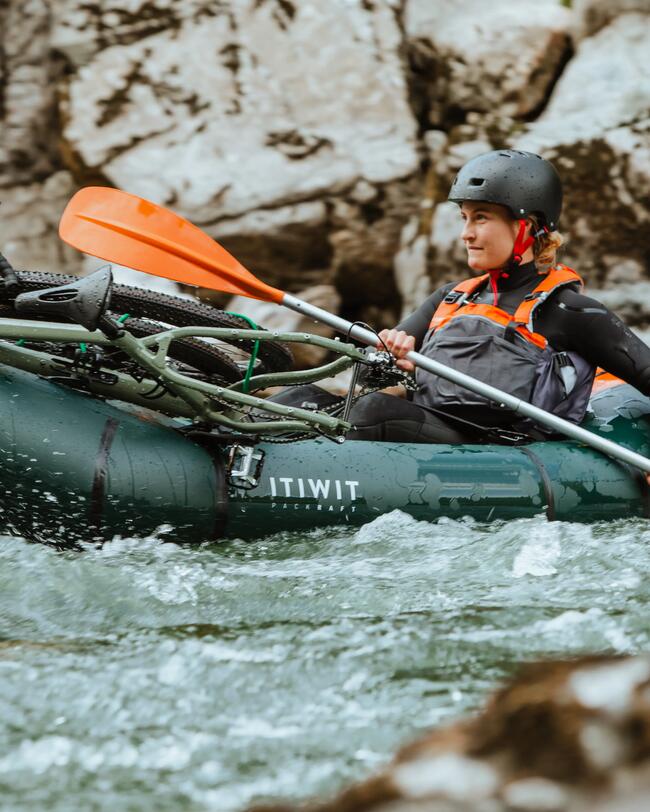 Person im grünen Packraft auf wildem, sprudelndem Wasser, orange Paddel, grauer Helm, felsiger Flussufer-Hintergrund.