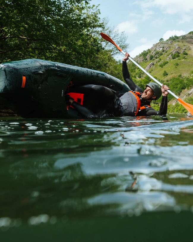 Person im Packraft, grünes Wasser, orange Paddel, grüne Uferbäume, Felsen im Hintergrund, heller Himmel, Outdoor-Abenteuer.