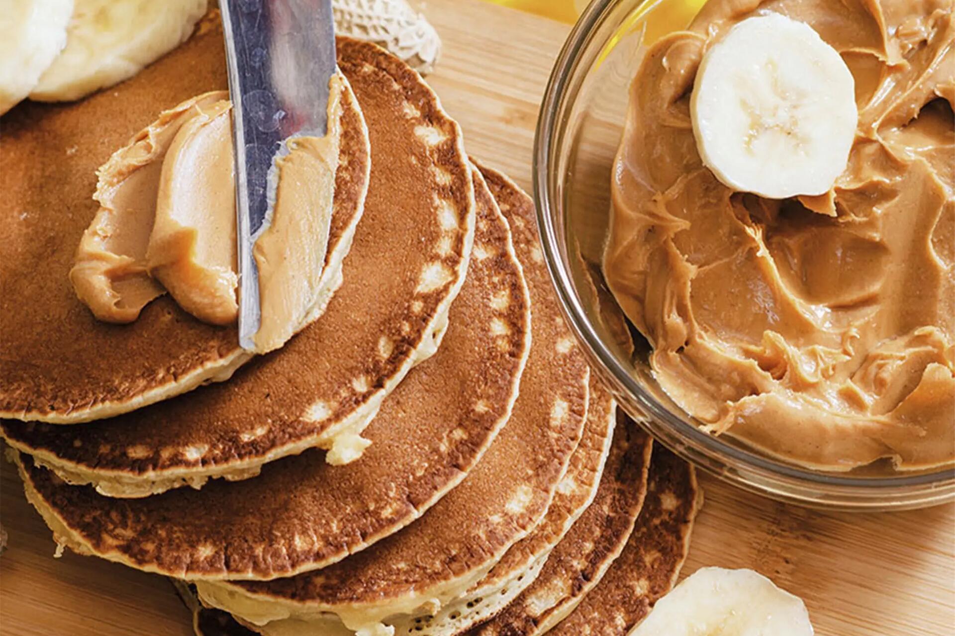 Nóż smarujący pancakes obok miseczka z masłem orzechowym i pokrojonymi bananami