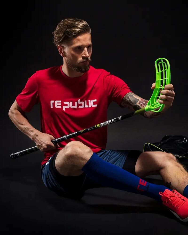 Ein Unihockeyspieler sitzt in Sportbekleidung auf dem Boden und begutachtet die neongrüne Kelle seines Schlägers, während neben ihm eine Sporttasche und ein Lochball liegen.