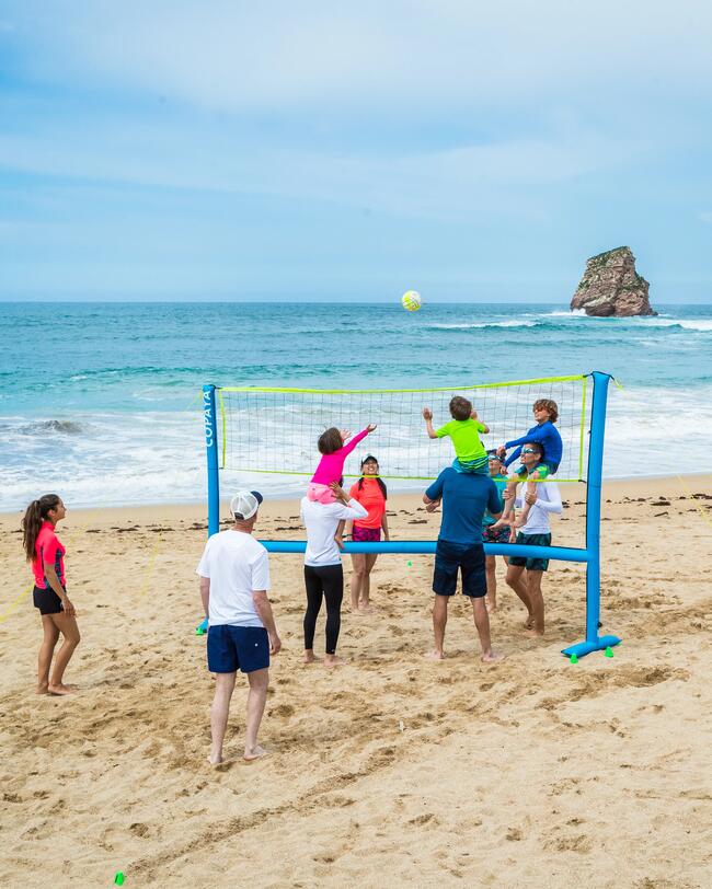 Personen spielen Volleyball am sandigen Strand mit blauem Meer, grünem Hügel und leichter Wolkendecke im Hintergrund.