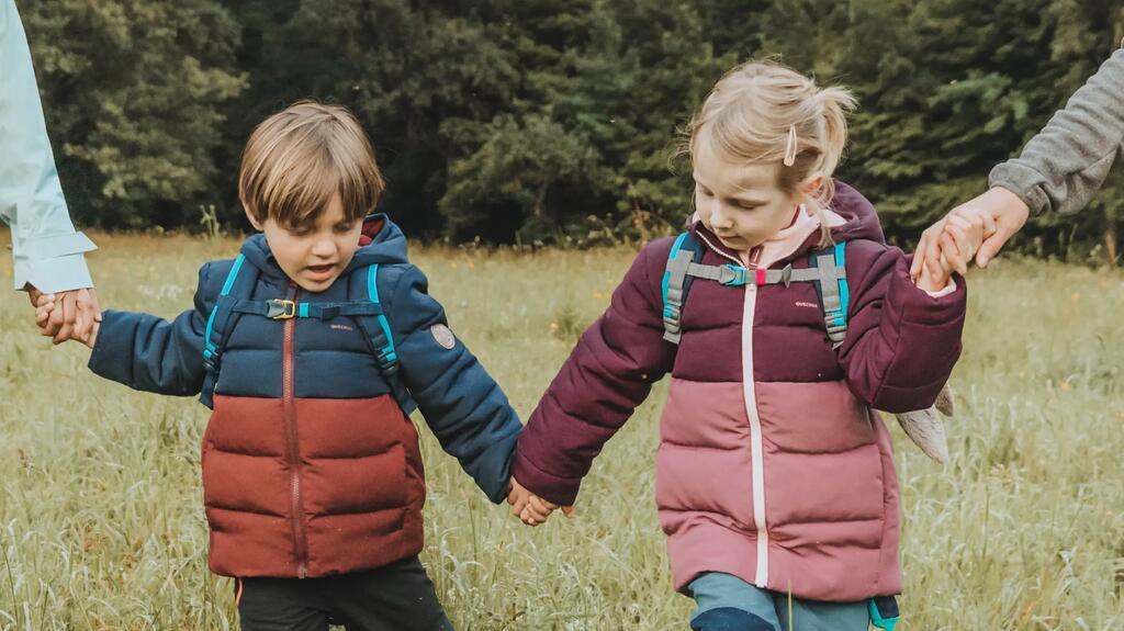 Zwei kleine Kinder in bunten Jacken gehen Hand in Hand über grüne Wiese vor bewaldeter Felswand.