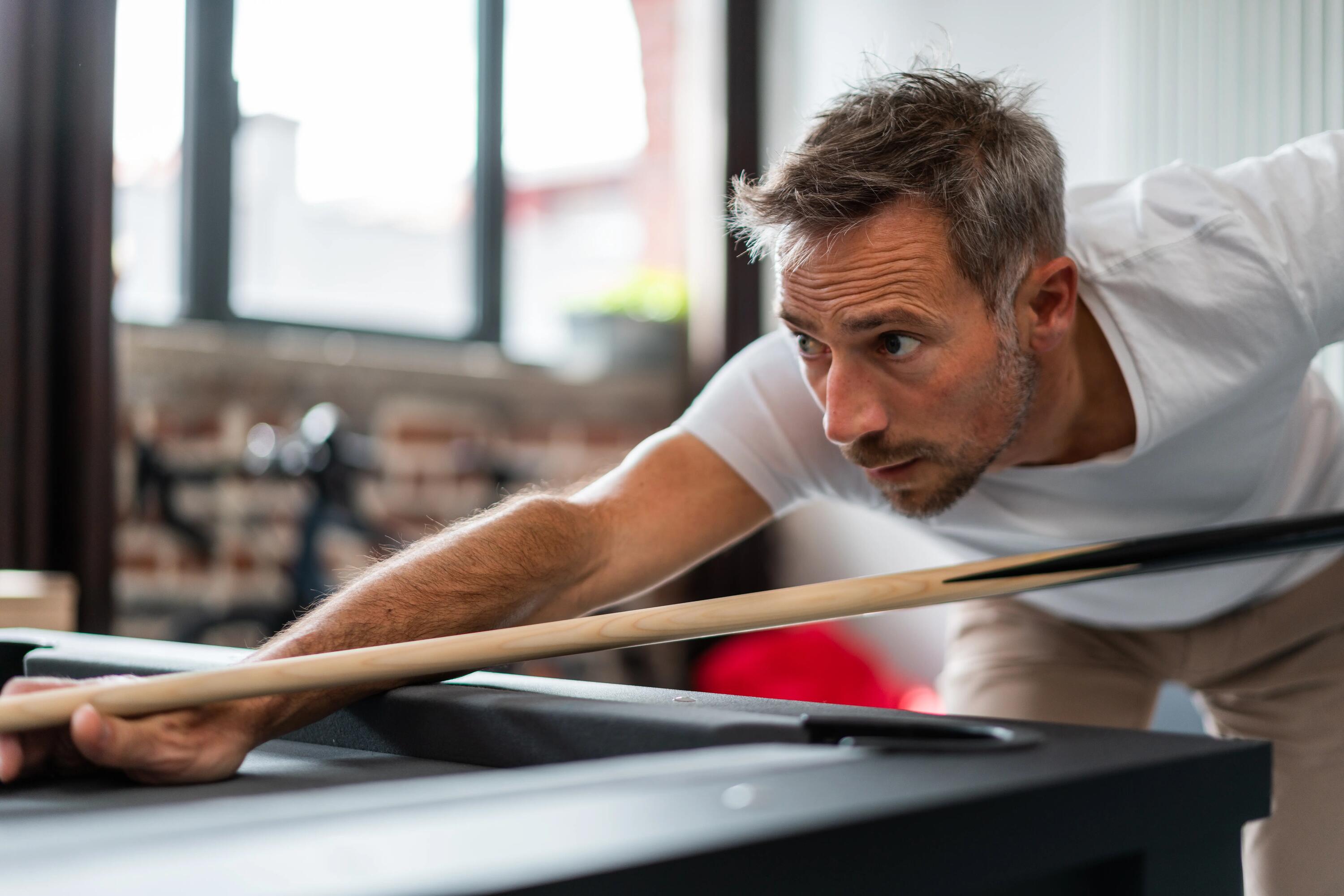 Fokussierter Pool-Billard-Spieler im weißen T-Shirt zielt mit einem Queue auf eine weiße Kugel auf einem modernen schwarzen Billardtisch; professionelles Billardzubehör wie Master's Kreide, Cue Oil und ein Pflegetool liegen griffbereit auf dem Filz.