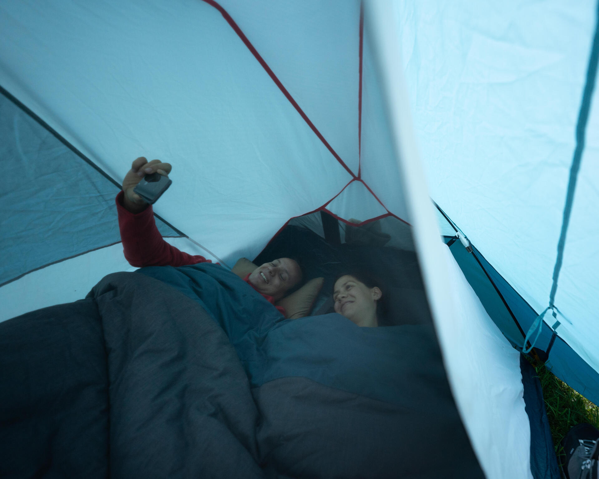 Deux campeuses sont confortablement installées dans leur tente et sur leur matelas de bivouac et se prennent en photo en souvenir avec un téléphone