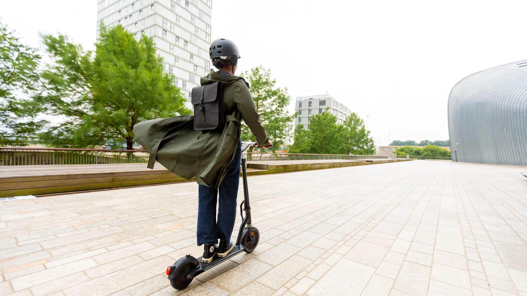 Frau auf einem E-Roller und einem Fahrradhelm in der Stadt unterwegs mit Rucksack auf dem Rücken.