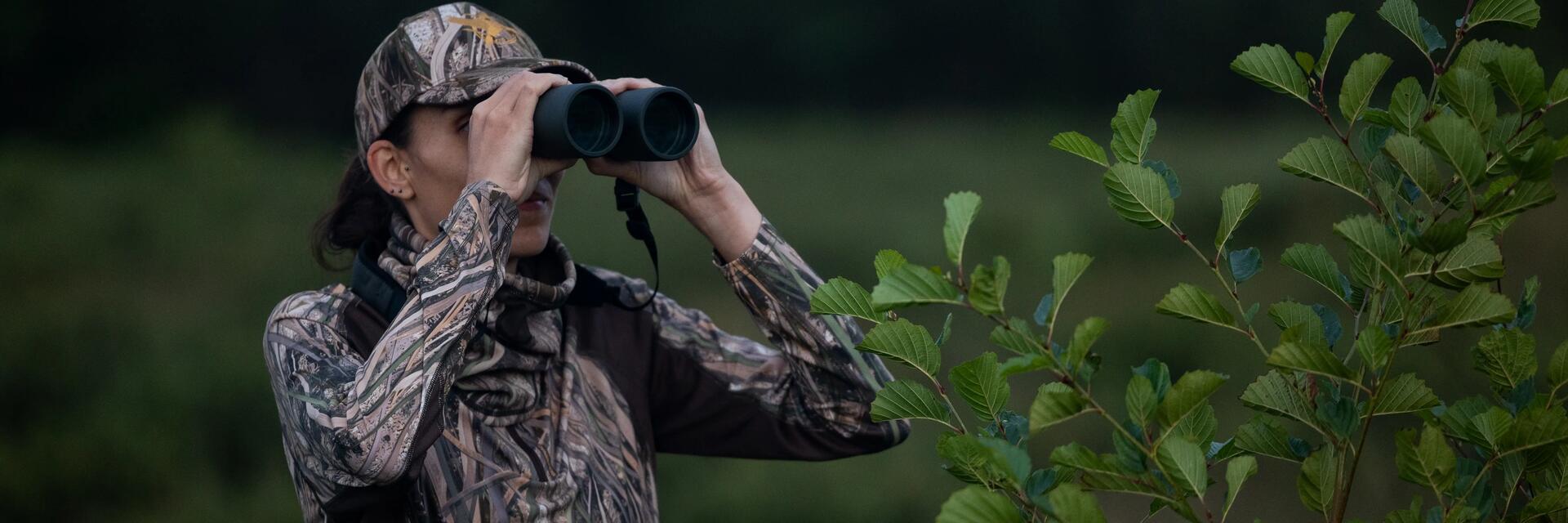 Kobieta w odzieży myśliwskiej patrząca przez lornętkę za krzaka