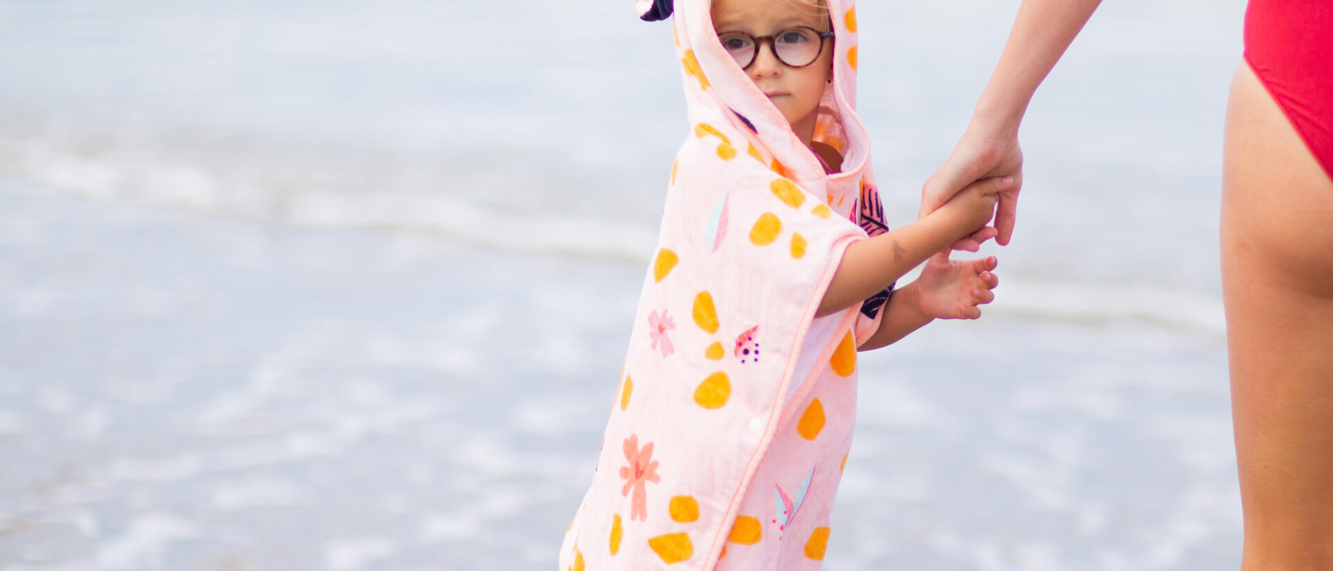 Criança com poncho Nabaiji rosa, junto à agua na praia de mão dada com uma mulher