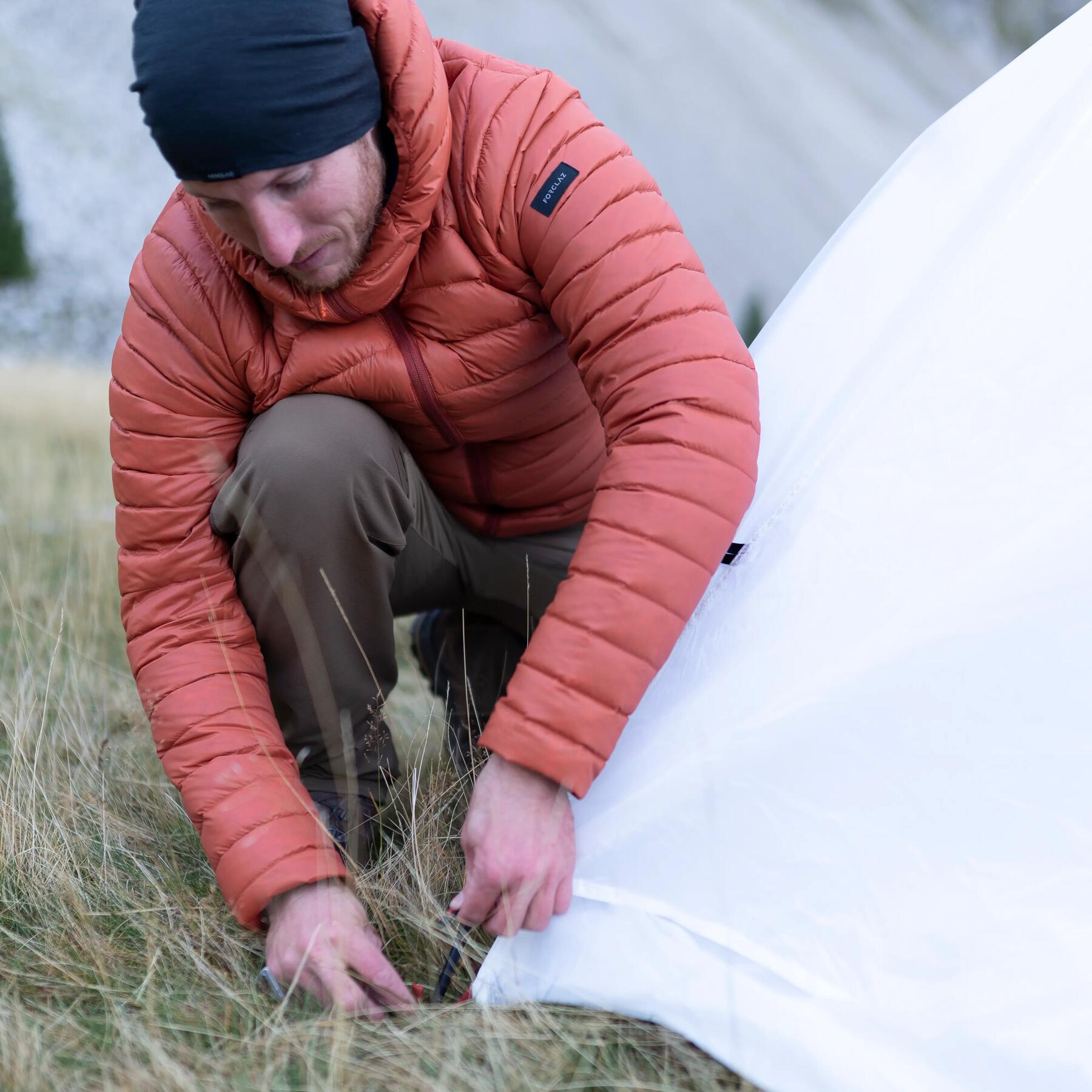 Mężczyzna w czapce i kurtce turystycznej wbijający śledzia namiotowego w ziemię obok rozłożonego namiotu
