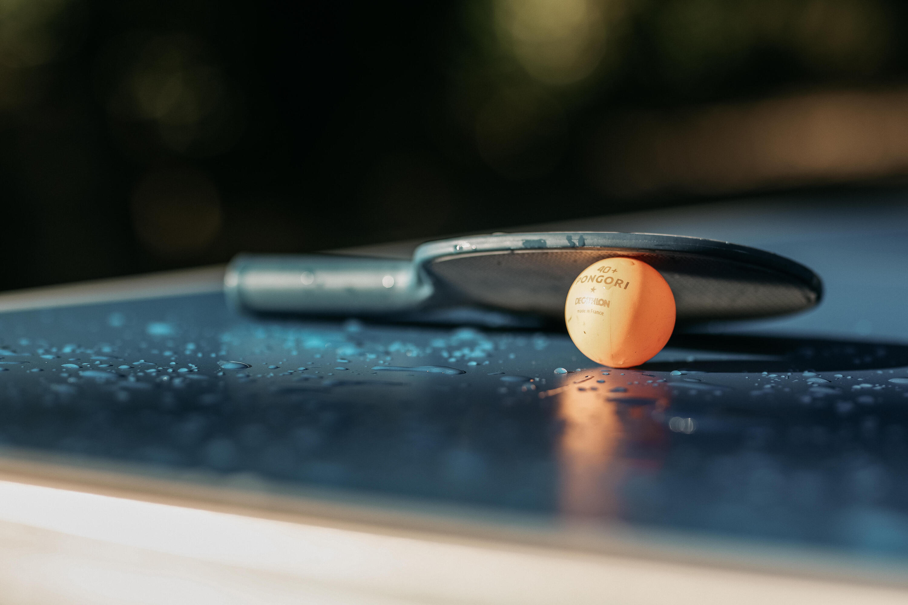 Ein Tischtennisschläger und ein Tischtennisball liegen auf einem nassen Tischtennistisch.