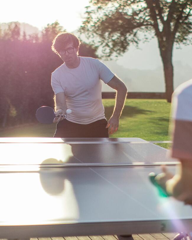 Zwei Männer spielen Tischtennis im Freien. Garten, Hecke, Baum und Berg im Hintergrund. Sonne scheint auf Tischtennisplatte.
