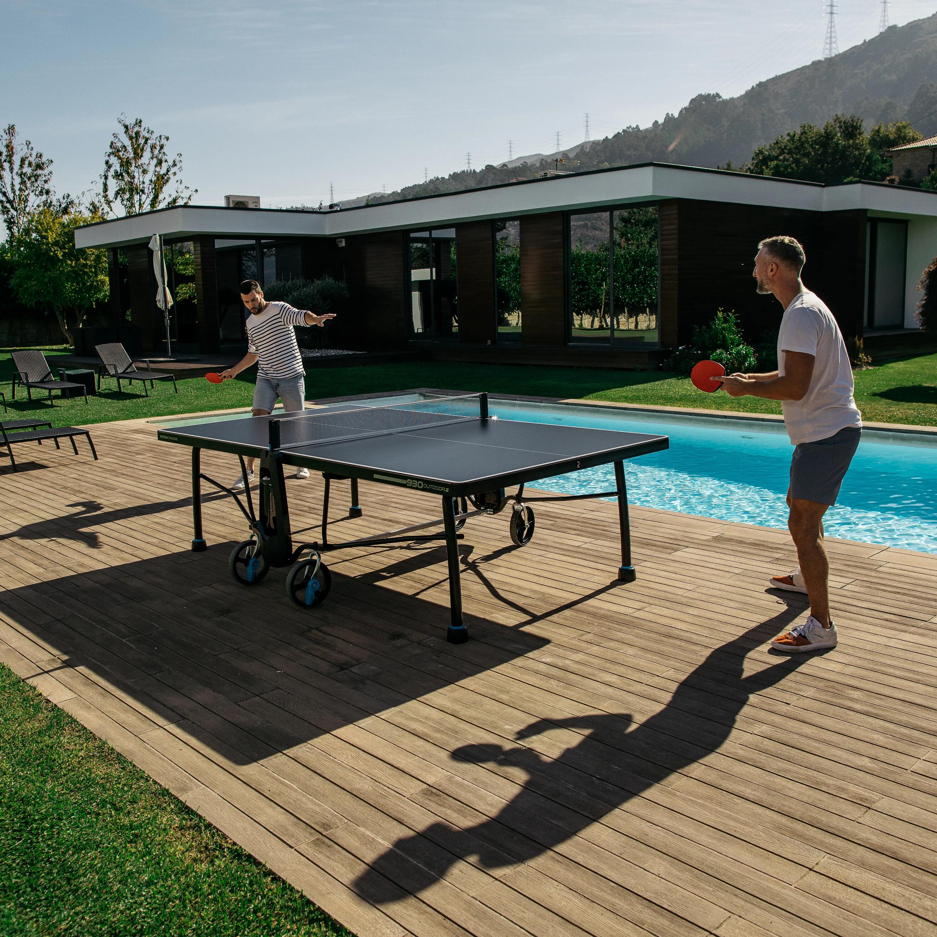 Zwei Männer spielen Tischtennis auf einer Holzterasse. Pool und Haus im Hintergrund.