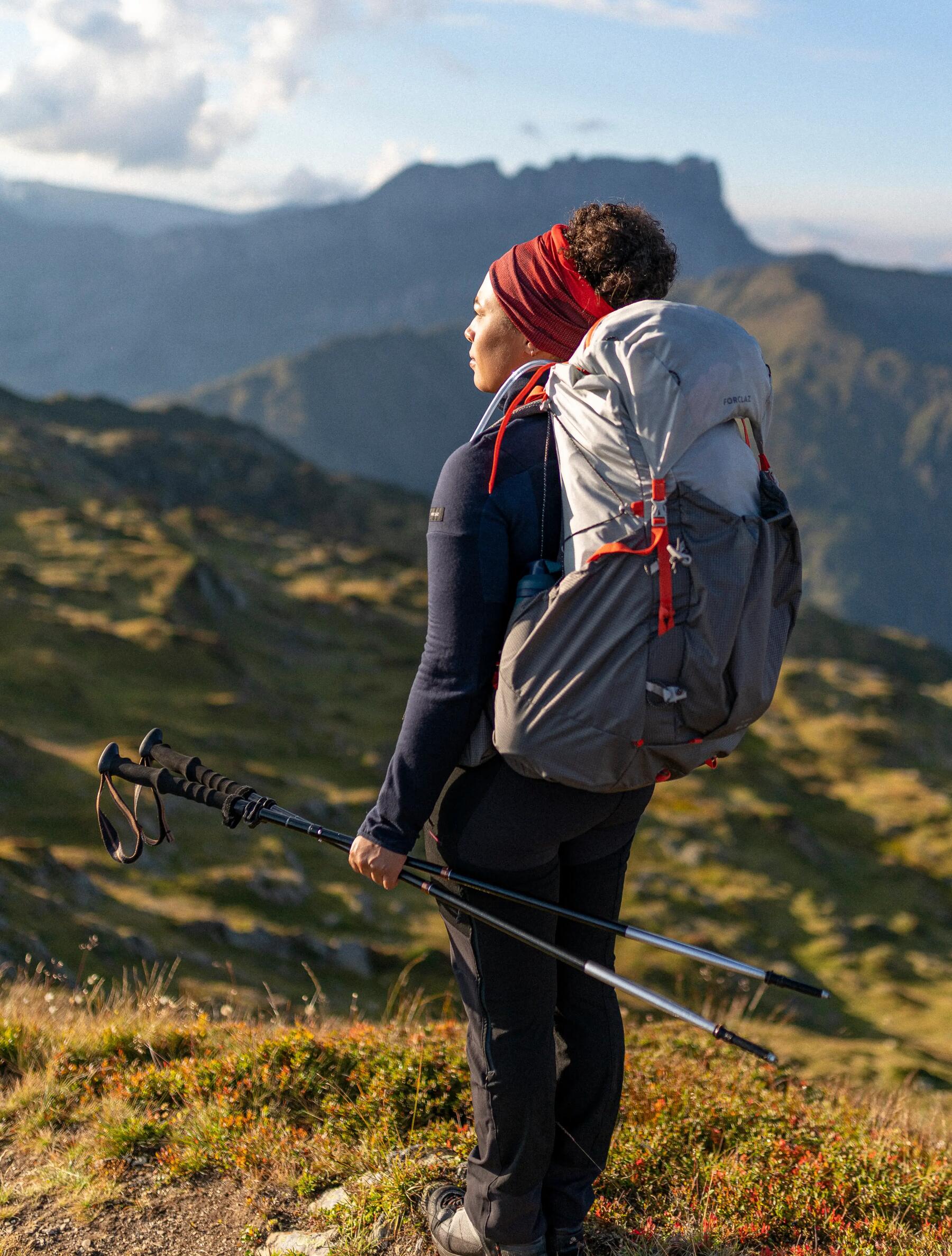 Kobieta w odzieży trekkingowej stojąca w górach patrząc w słońce z kijami trekkingowymi w rękach