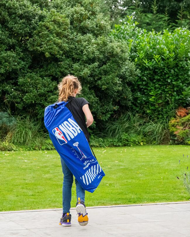 Kind trägt blauen Tragetasche für tragbaren Basketballkorb im Garten, leichter Transport, grüne Büsche im Hintergrund.
