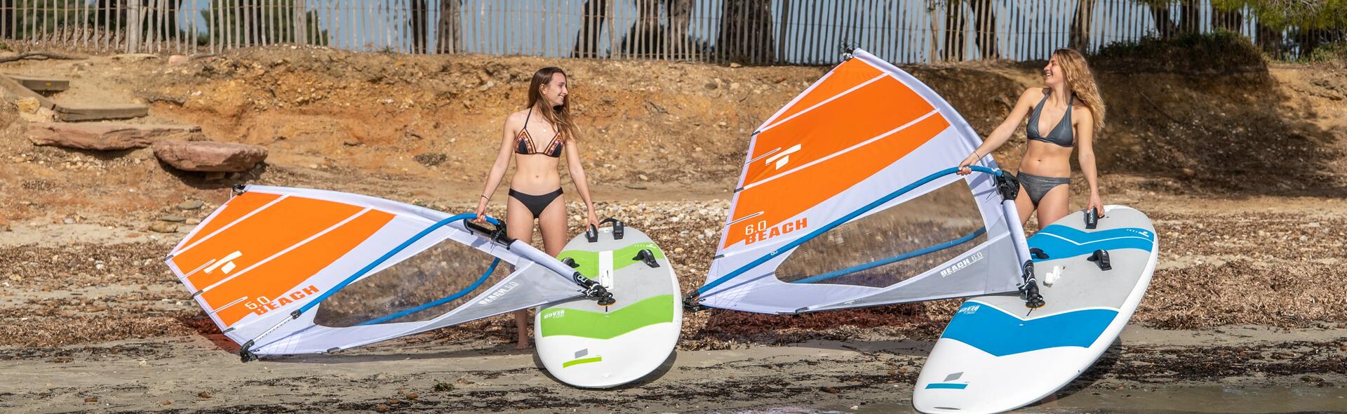 kobiety w strojach kąpielowych stojące na brzegu trzymając deski windsurfingowe z żaglami