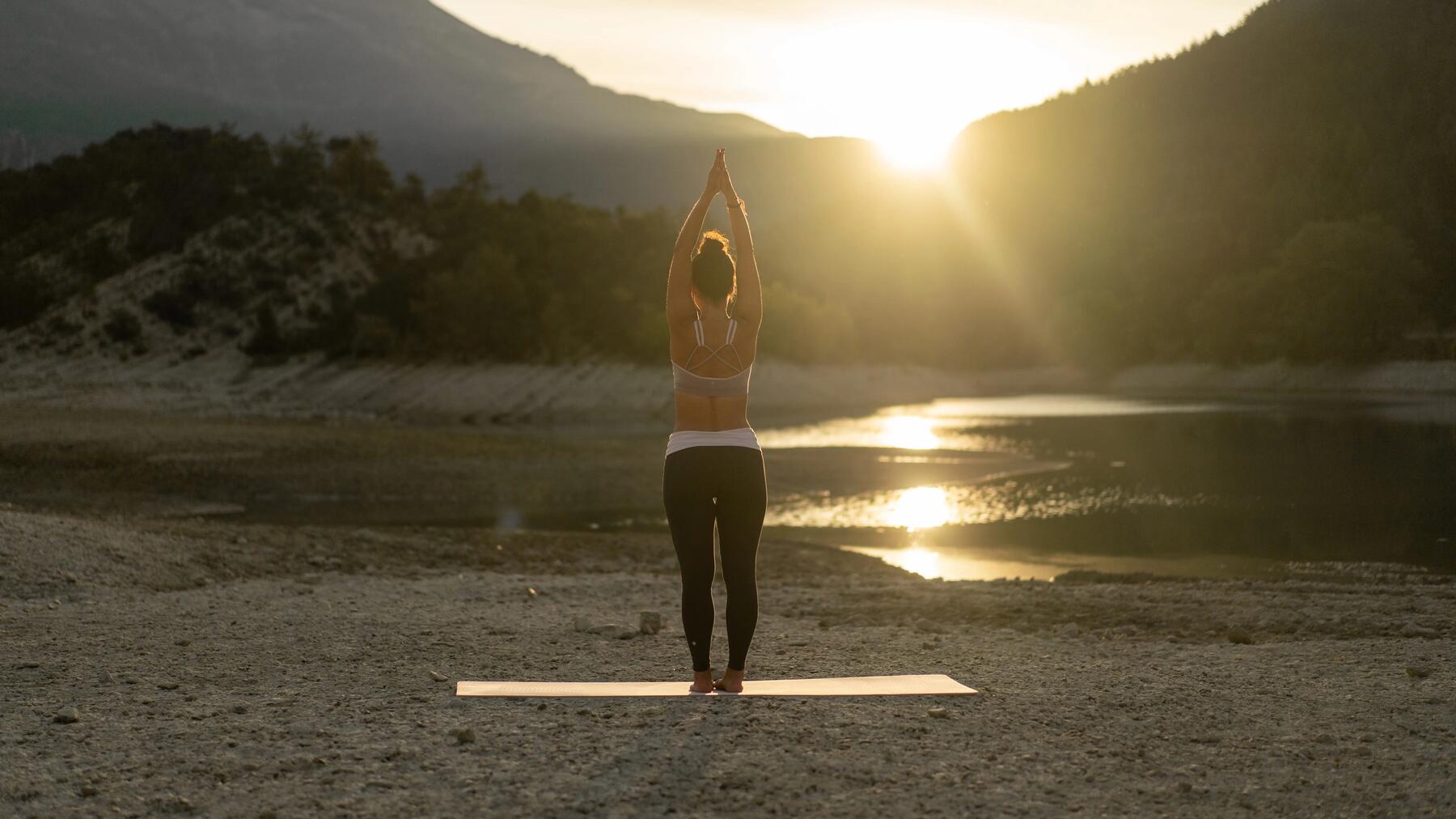 Frau steht auf ihrer Yogamatte auf Steinen vor See bei Sonnenaufgang.
