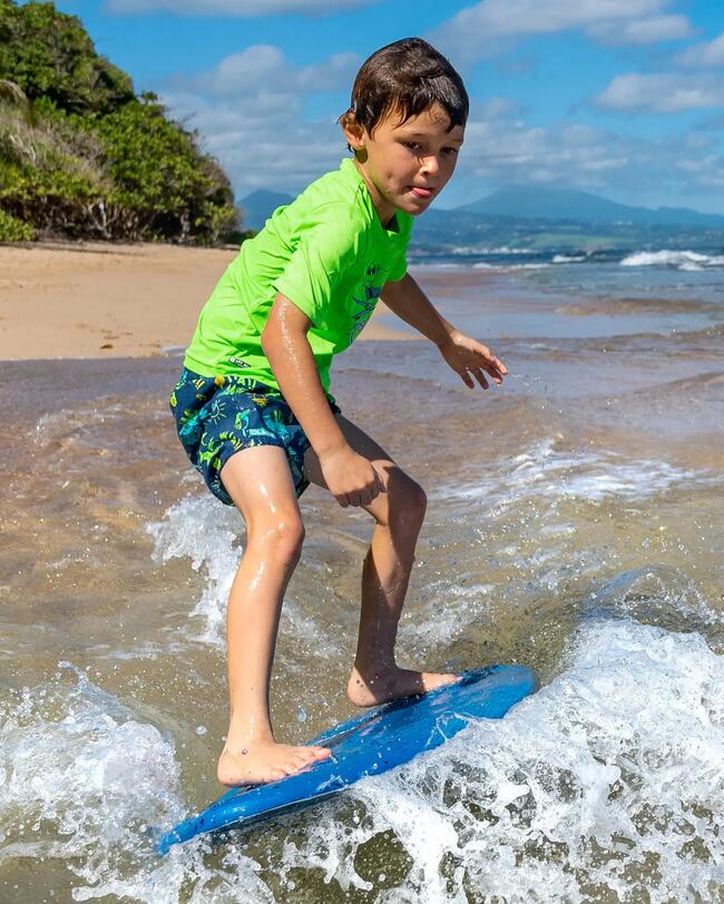 Kind surft auf blauem Brett im Meer; trägt grünes Shirt und blaue Shorts; Wellen und Sandstrand im Hintergrund; sonniger Himmel.