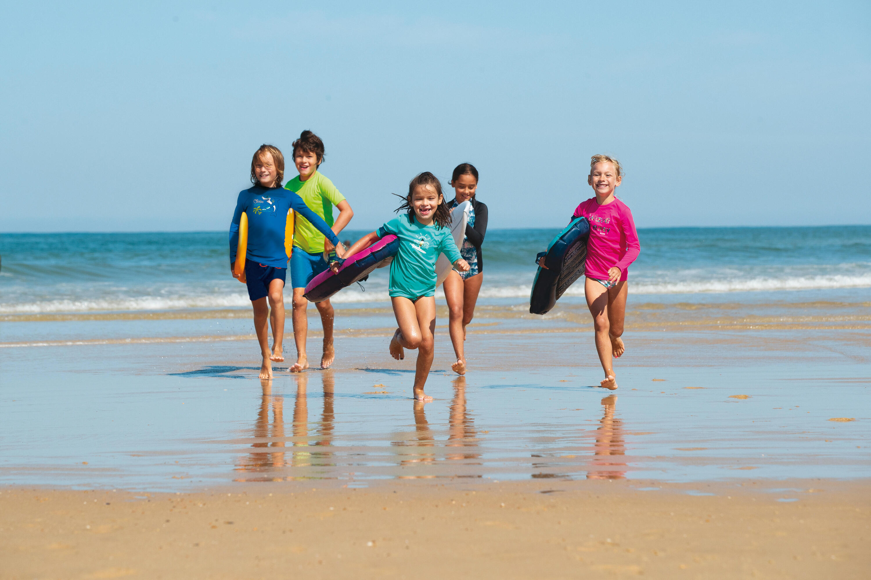 Kinder in bunten Badesachen rennen lachend am Sandstrand entlang, Wellen im Hintergrund, strahlend blauer Himmel.