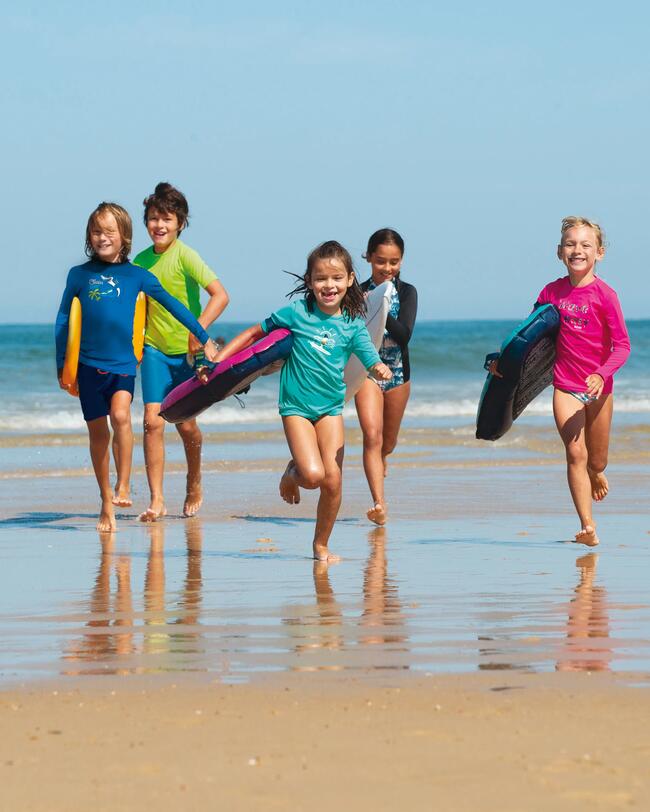 Kinder laufen am Strand, bunte Surfbretter, blaues Meer, klare Himmel, nasser Sand, lachende Gesichter, strahlende Farben.