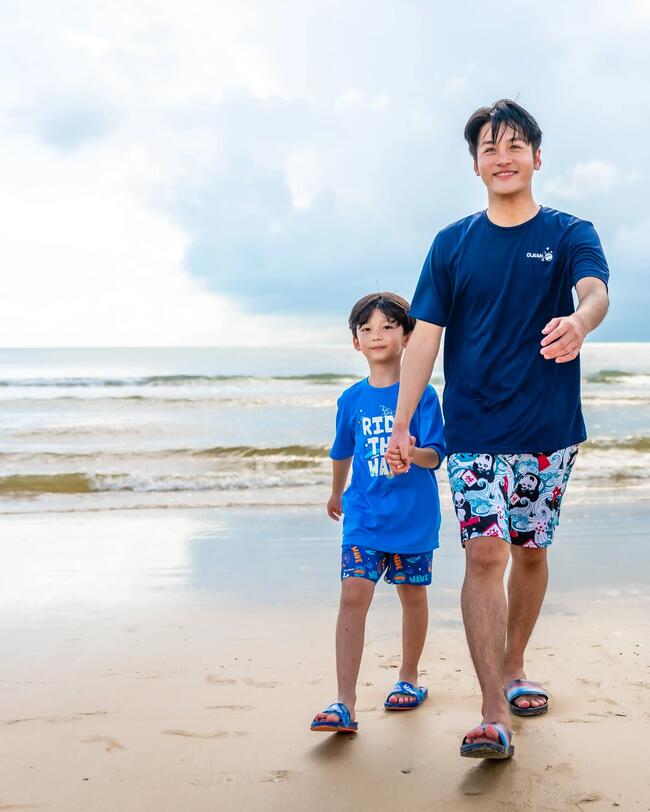 Zwei Personen am Strand in blauen Shirts und bunter Badehose, nasser Sand, wolkiger Himmel, sanfte Wellen im Hintergrund.