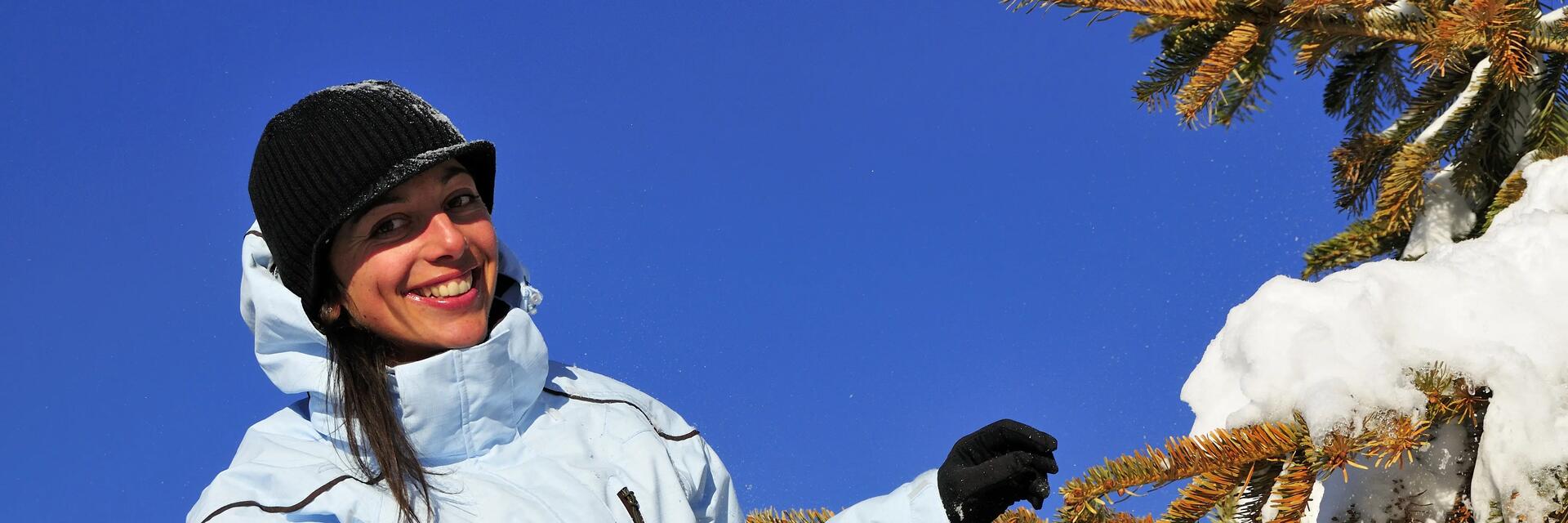 Frau in weißer Winterjacke und schwarzer Mütze neben Nadelbaum im Schnee.