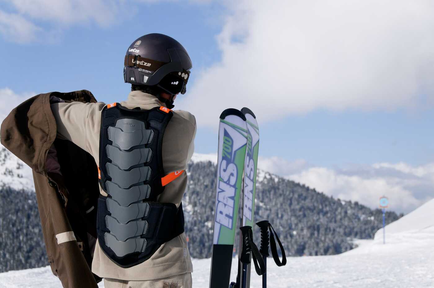 Skifahrer in Schutzausrüstung (Helm, Brille, Rückenprotektor) auf einem schneebedeckten Berg, der seine Skijacke anzieht.