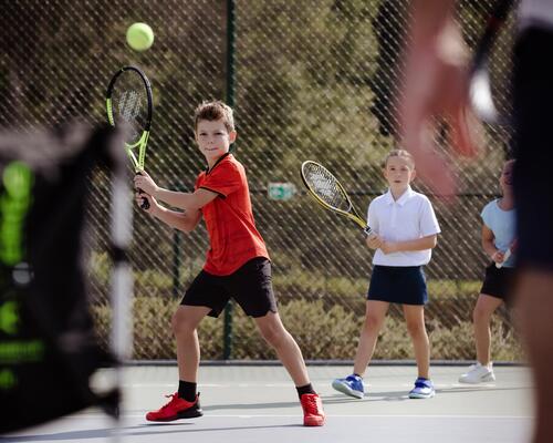 Ein Junge in einem roten Shirt holt auf einem Tennisplatz zum Schlag aus, beobachtet von zwei Mädchen im Hintergrund und einem Trainer im Vordergrund