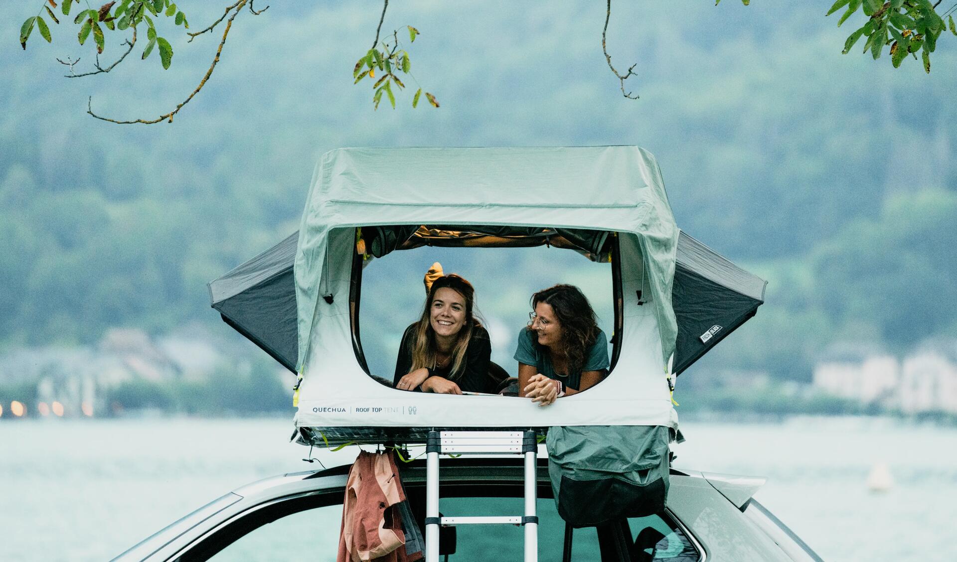 Zwei Personen liegen in einem Dachzelt und schauen in die Natur, mit einem See im Hintergund.