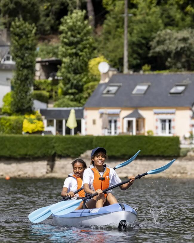 Zwei Personen im blauen Kajak auf Wasser, tragen orange Westen; grüner Hintergrund mit Bäumen und Haus.