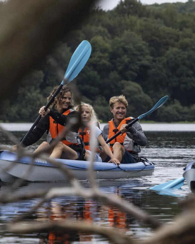 Drei Personen in blauen Kajaks, tragen orange Schwimmwesten, paddeln durch ruhiges Wasser, umgeben von grünen Bäumen.