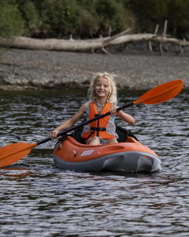Kind in orangefarbenem Hartschalenkajak auf ruhigem Wasser, grünes Ufer im Hintergrund, lachend, mit Paddel, Sommeratmosphäre.