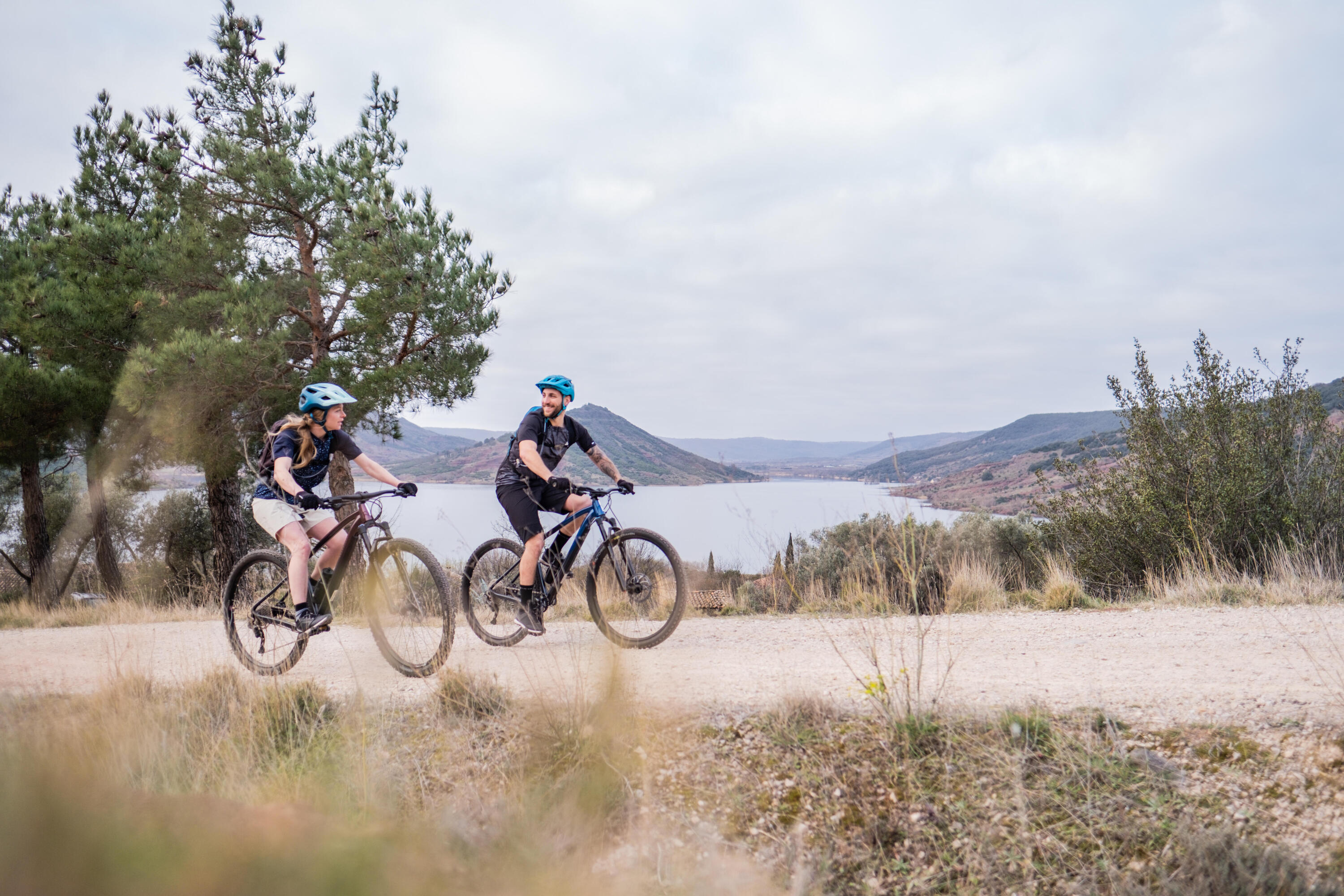 zwei Personen auf Mountainbikes, die nebeneinander auf einem unbefestigten Weg fahren. Im Hintergrund erstreckt sich eine Hügellandschaft mit einem großen See unter einem bewölkten Himmel.