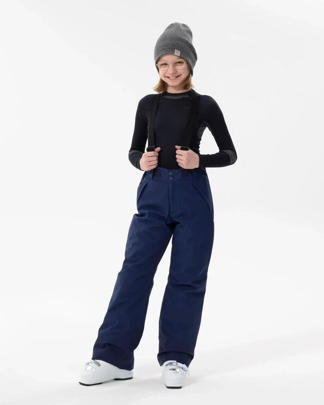Ein Mädchen mit schwarzem Baselayer und grauer Mütze trägt die dunkelblaue Skihose mit Hosenträgern und lacht dabei in die Kamera.