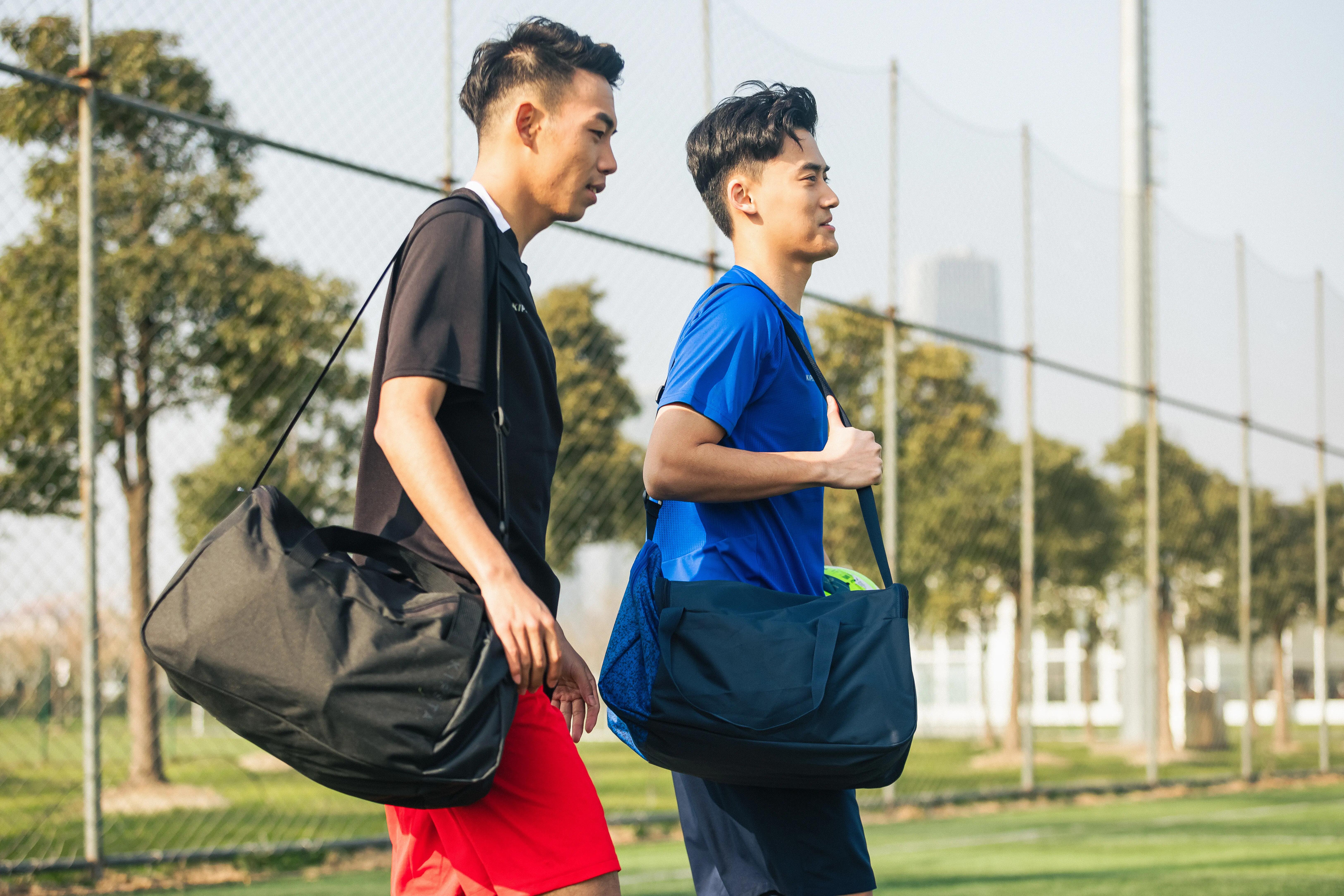 Two athletes carrying large sports bags while walking beside an outdoor sports court.