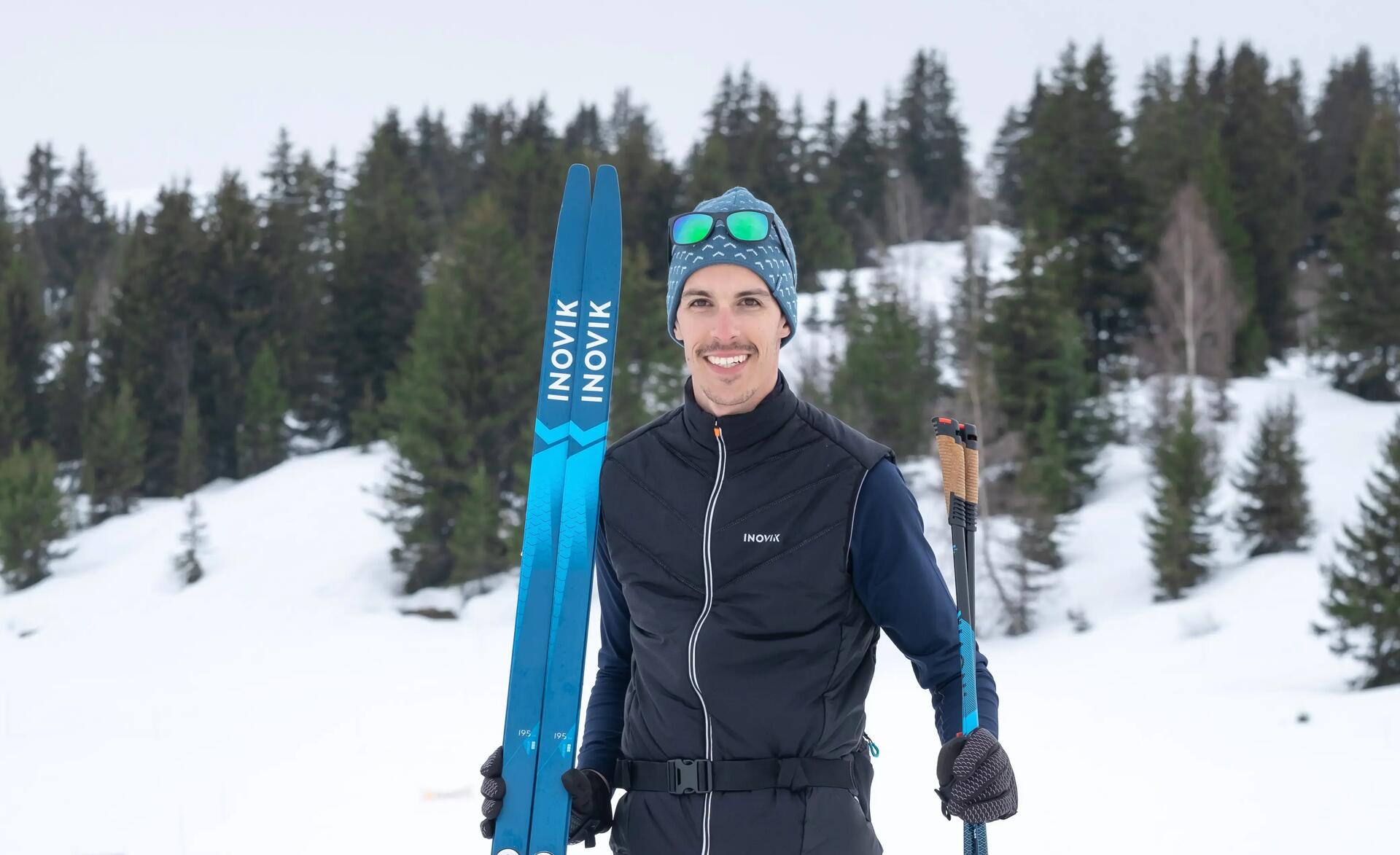mężczyzna w odzieży narciarskiej trzymający narty biegowe i kijki narciarskie 