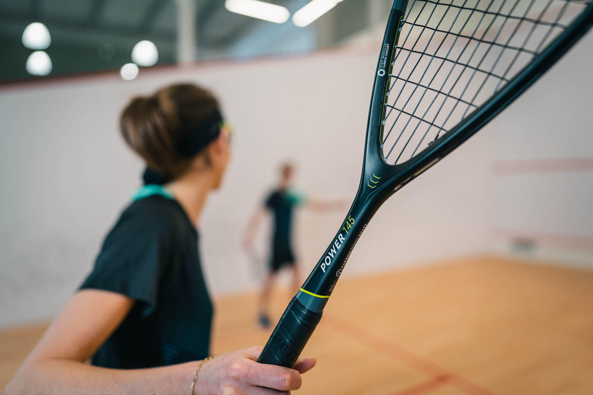 Black squash racquet held close-up, players on court behind