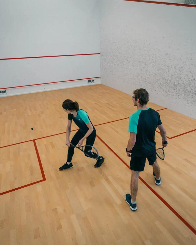 Eine junge Frau und ein Mann stehen auf einem Squash-Court mit Holzboden und weißen Wänden; die Spielerin ist gerade dabei, den Ball mit ihrem Schläger tief anzunehmen.
