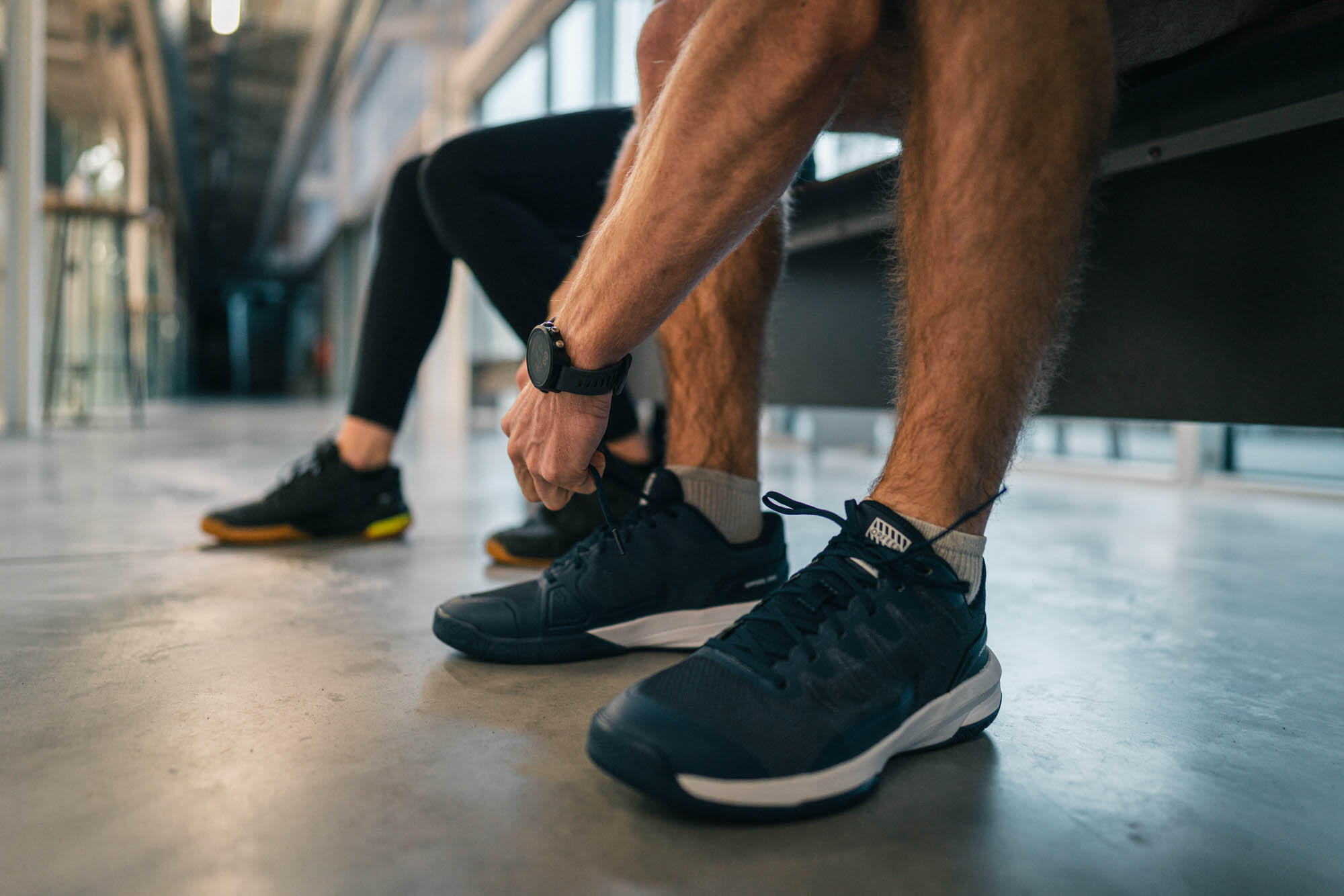 Nahaufnahme der Beine und Füße eines Mannes, der auf einer Bank sitzt und sich die dunklen Sportschuhe bindet.