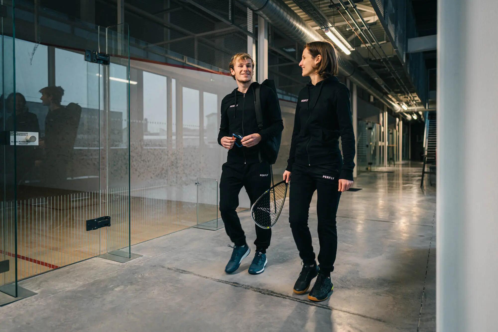 Ein Mann und eine Frau, beide in schwarzen Perfly-Anzügen, gehen gut gelaunt einen Flur mit Squash-Courts entlang. Der Mann trägt eine Sporttasche und die Frau einen Squash-Schläger. Beide lächeln sich an.