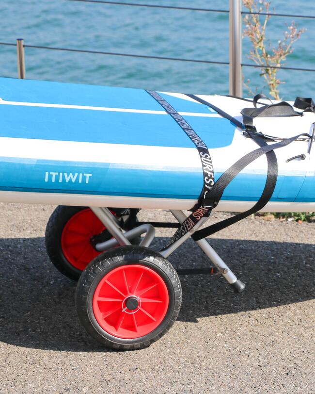 Blaues Paddelbrett auf einem Wagen mit roten Rädern, befestigt mit schwarzen Gurten, im Hintergrund ein See und Geländer.