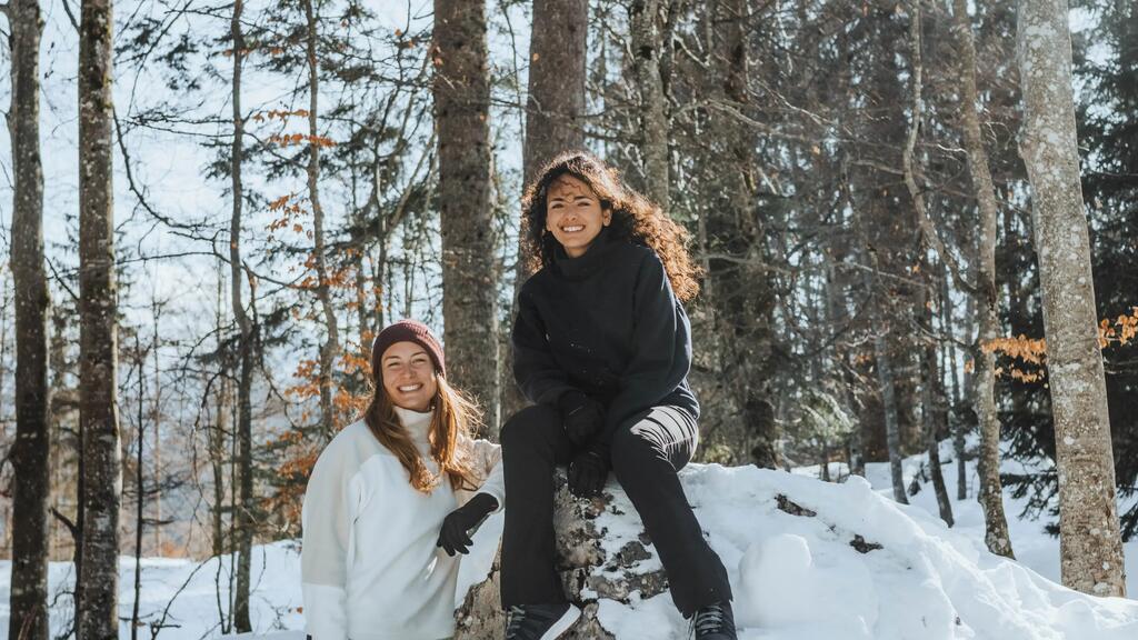 Zwei Frauen mit dunkler Kleidung und weißen Pullovern vor verschneiten Bäumen, sitzen und stehen auf einem schneebedeckten Felsen.