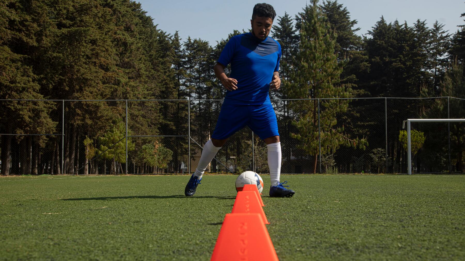 Mann in blauem kurzem Trainingsoutfit trainiert Dribbling mit Kegeln. Bäume im Hintergrund.