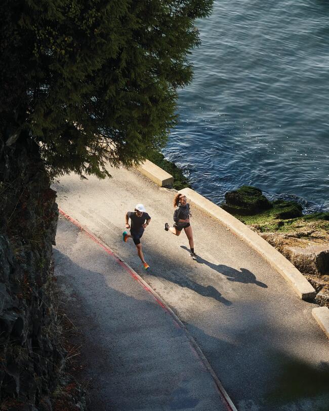 Ein Mann und eine Frau joggen am Wasser entlang. Aufnahme aus der Vogelperspektive.
