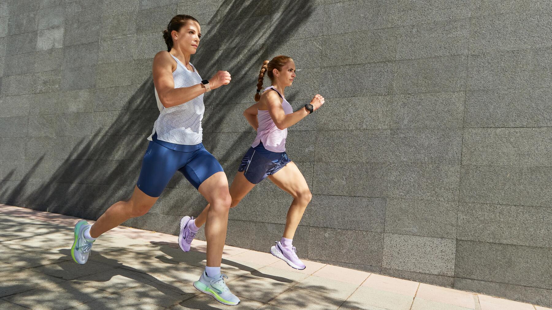 Zwei Frauen laufen sportlich an einer grauen Steinwand entlang, tragen Sportkleidung in Blau, Lila und Weiß, klarer Schattenfall.
