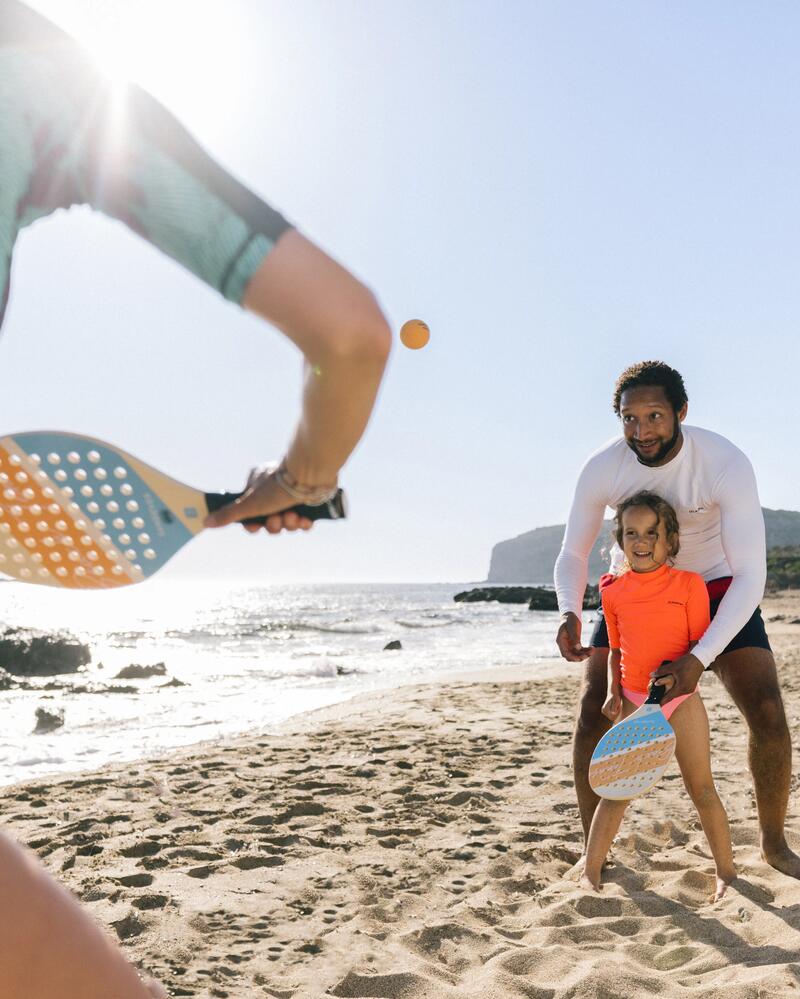Ein Vater hilft seiner Tochter am sonnigen Strand dabei, den Beach-Tennis-Schläger richtig zu halten, während ihnen eine weitere Person einen Ball zuspielt.