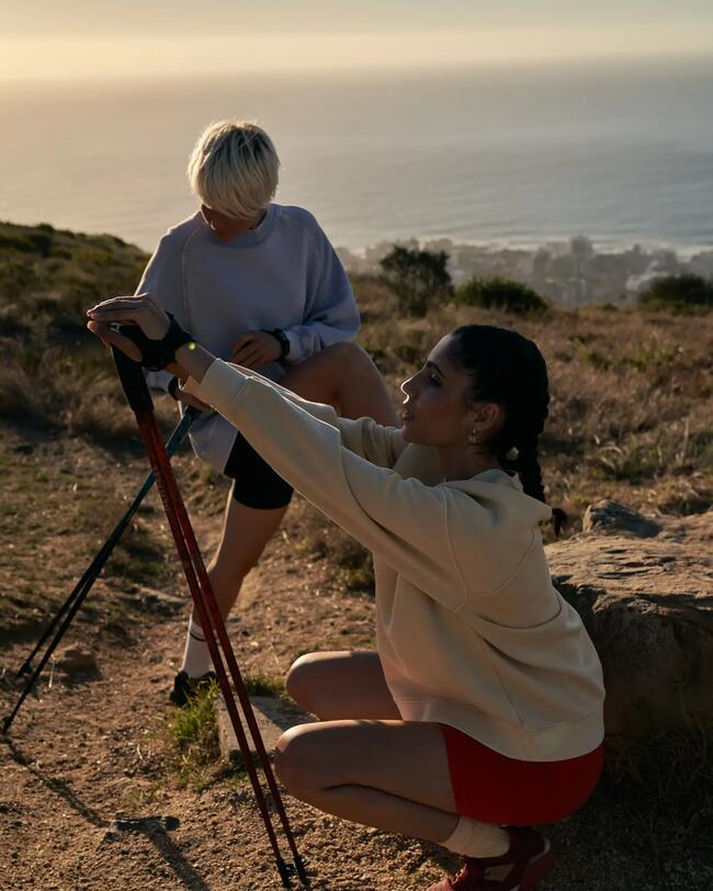 Zwei Personen beim Wandern, eine hockt mit Stöcken, die andere steht. Sonnenuntergang, Meer und Hügel im Hintergrund.