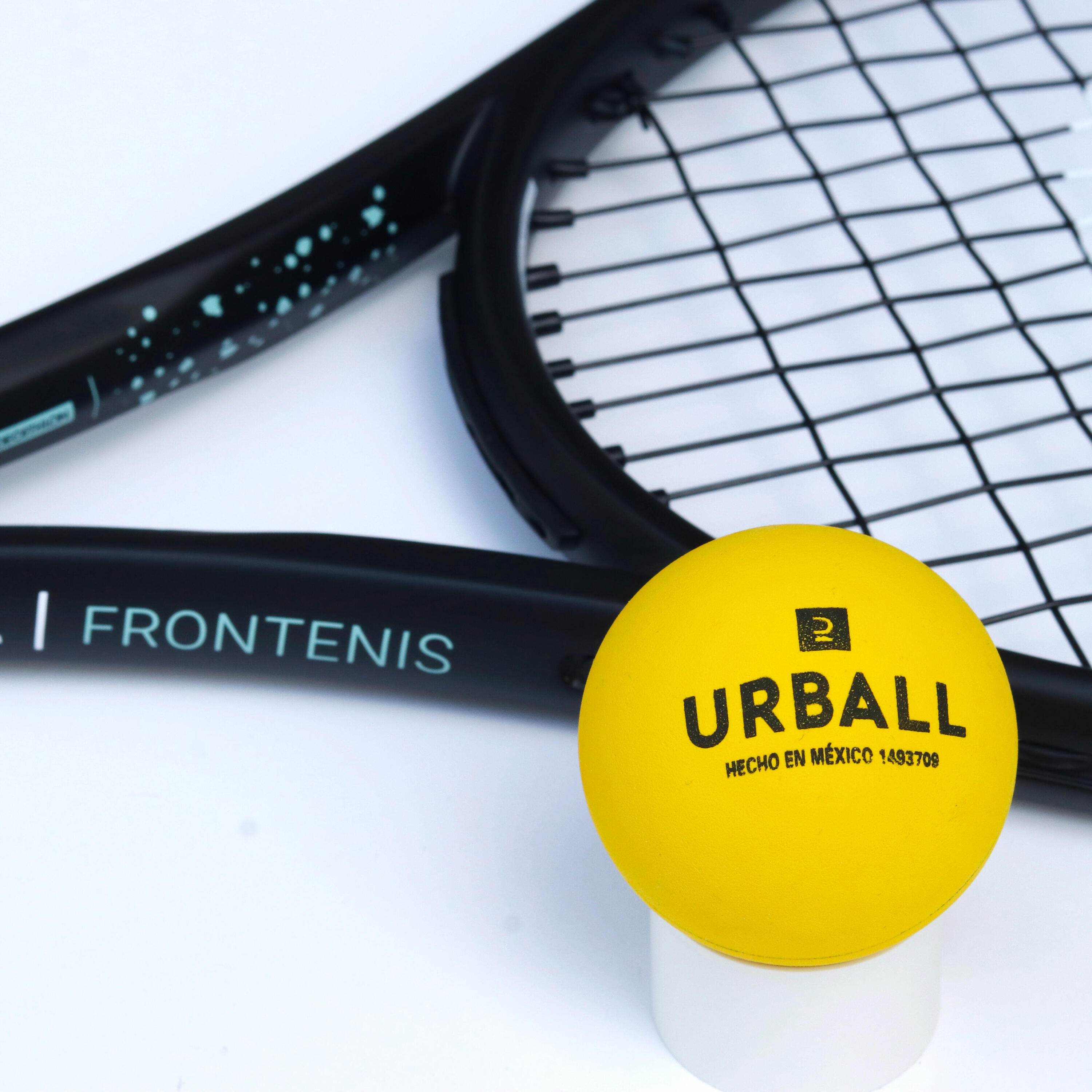 Alt-Text: Nahaufnahme eines gelben Urball-Frontenis-Balls vor einem schwarzen Frontenis-Schläger mit türkisblauem Schriftzug und Pünktchen-Muster vor weißem Hintergrund.