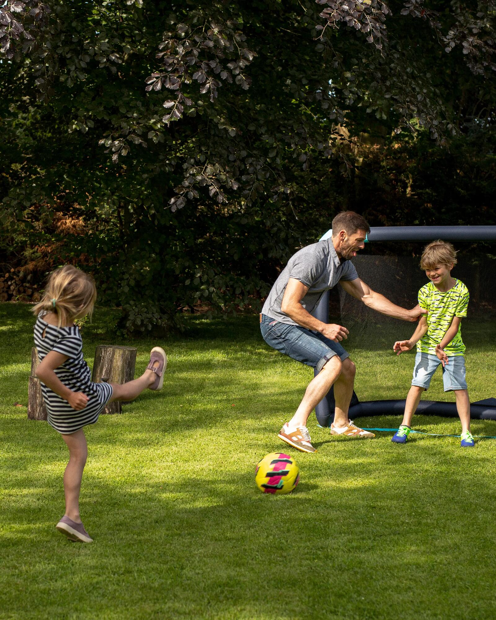 Vater und zwei Kinder spielen Fußball im Garten mit einem aufblasbaren Tor auf einer grünen Wiese.