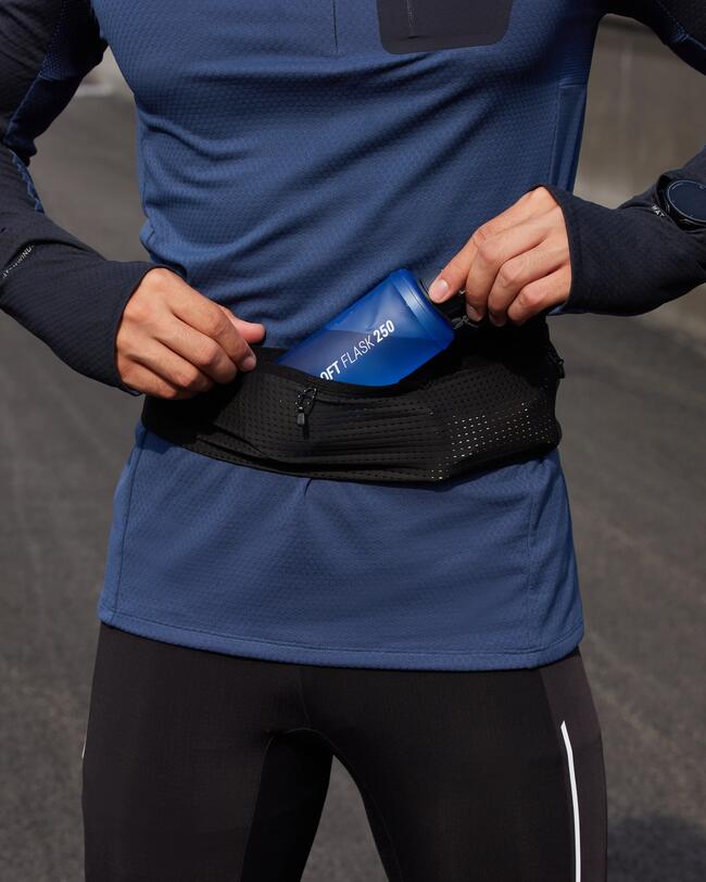 Person in blauem Langarmshirt und schwarzen Leggings steckt blaue Flasche in schwarzen Gürtel vor asphaltiertem Hintergrund.